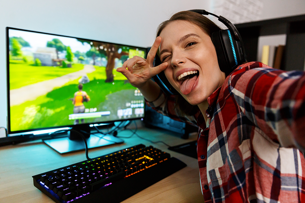 Female web streamer in a checked shirt poses with a victory symbol, happy sticking out her tounge whilst a game is playing on her computer in the background.