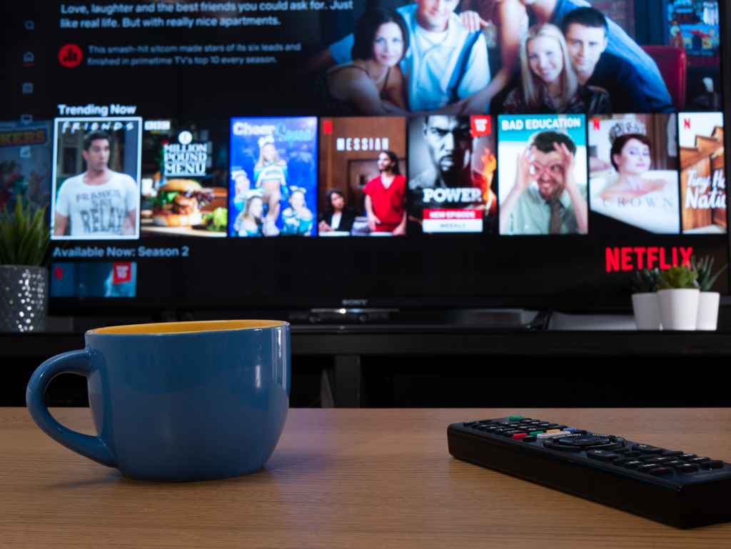 Netflix menu is shown on the TV which fills the background. In the foreground, we see a coffe table with a big cup of coffee in a blue cup and a TV remote control.  Image to illustrate that VoIP won't affect streaming services.