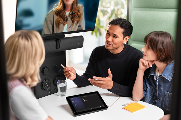 Close-up image of a conference room featuring jabra Pancast equipment, three meeting room users and one remote user on a view screen. We can see a table, a control panel, a Pancast camera and speaker bar and some notepads and pens on the table.