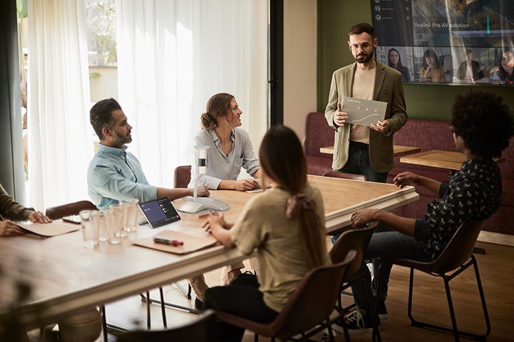 A boardroom scene featuring six  people using Yealink Conferencing Equipment to meet with their colleagues we can see on a large screen in the background.